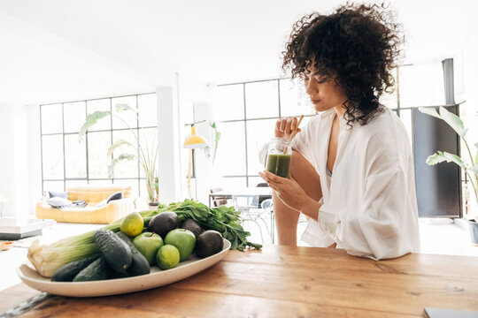 Young african american pretty woman relaxing in kitchen while having a green juice with bamboo straw in the morning. Detox. Cleanse.