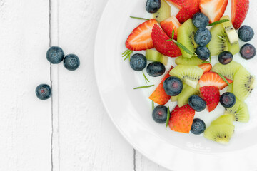 a flatlay shot of ripe strawberries, kiwi fruit, blueberries, yoghurt and honey on a white plate, shot on a textured background