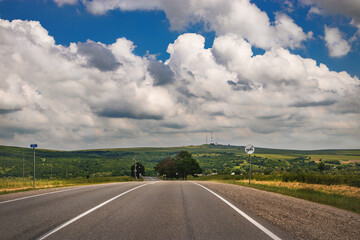 An empty expressway through a field of green grass and a blue sky with white cumulus clouds.