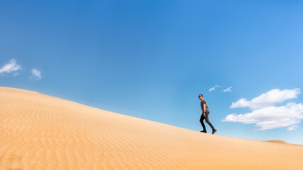 Fototapeta na wymiar A man climbs the dunes of the desert. The guy walks on the sand. Sunny day blue sky.