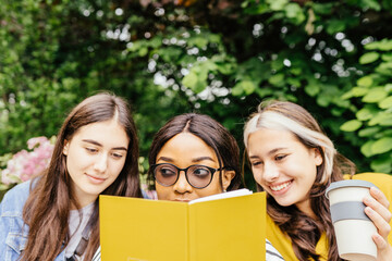 Three female students reading books together outdoor. University female friends of different nationalities sitting on the grass at the park in the morning reading a paperback book together.