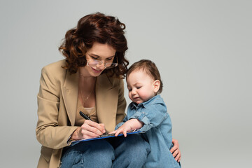 smiling businesswoman in glasses writing on clipboard near toddler son isolated on grey