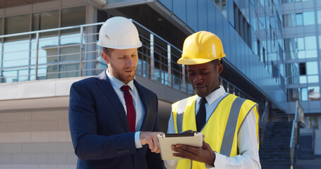 Diverse businessman and architect in hardhats with tablet pc computer at construction site