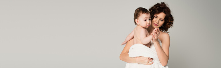 caring mother with bare shoulders holding hands with baby boy wrapped in towel isolated on grey, banner