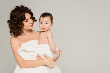 mother with bare shoulders holding in arms baby boy in towel isolated on grey