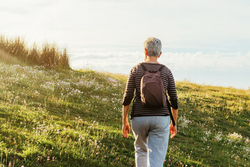 Retired woman with gray hair with backpack taking a walk in a natural setting at sunset. Old age and healthy lifestyle.