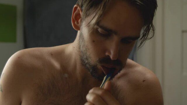 Bearded shirtless man in bathroom brushing his teeth with eyes closed