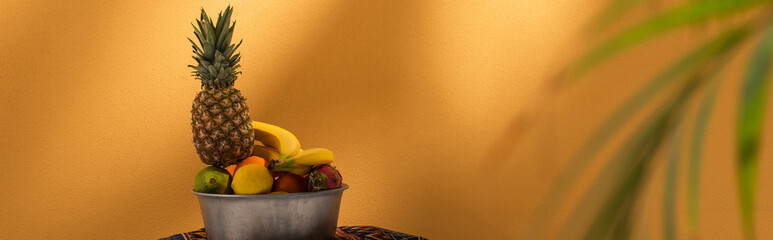 exotic fruits in metal bowl near blurred palm leaves on orange, banner