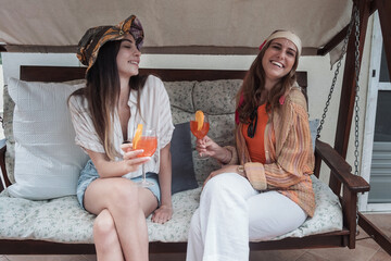 Obraz premium Cheerful young women talking together sitting in the rocking chair and drinking tropical fruit cocktails together