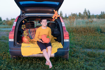 A cheerful and funny baby boy sits in the trunk of a car decorated with decor in honor of the autumn holiday Halloween, holding a decorative pumpkin of orange color. Lifestyle.