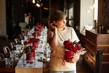 Caucasian girl florist decorator decorates the wedding table with red roses and eucalyptus in red boxes with red candles and chili and serves the table with glasses, plates and napkins