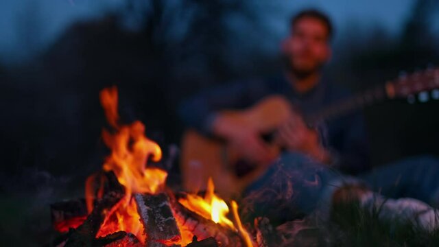 Evening at bonfire. Red and orange fire flame at night. Bright campfire on blur background of a man playing the guitar.