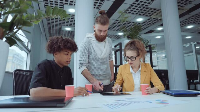 Startup team discussing progress together, comparing statistics on papers and tablet. Tracking shot cheerful multiethnic managers working in open space office. Concept of brainstorming