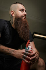 Portrait of a hipster in a gym. Concept of healthy lifestyle. Bearded tattooed man sitting and resting at a gym. 