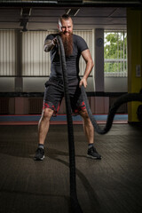 Strong young bearded man working out with battle ropes a gym. Determined guy using battle rope while doing physical training. Hard workout