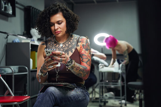 Serious woman with tattoos using phone in tattoo studio