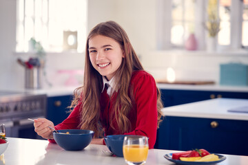 Portrait Of Smiling Teenage Girl Wearing School Uniform In Kitchen Eating Healthy Breakfast - obrazy, fototapety, plakaty
