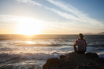 girl sitting in lotus position and meditating on a rock at sunset and looking at the ocean