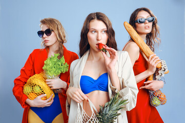 Three young fashionable females housewives in red and blue swimsuits and trench coats on naked body. Shopper strip bags with food in hands. Shopping, purchasing and consumerism. Beautiful long legs.