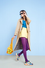 A young fashion girl in a beige trench, yellow short skirt, purple tights and sunglasses holds a mesh reusable eco bag with food.