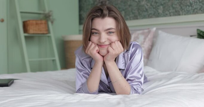 Young beautiful woman lies in pajamas on a large bed Bedroom of green gentle color Model holds her face with her hands and smiles Man happiness is resting alone on a soft mattress 
