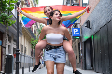 Two women smiling and piggyback riding with a lgbt flag. LGBT people.