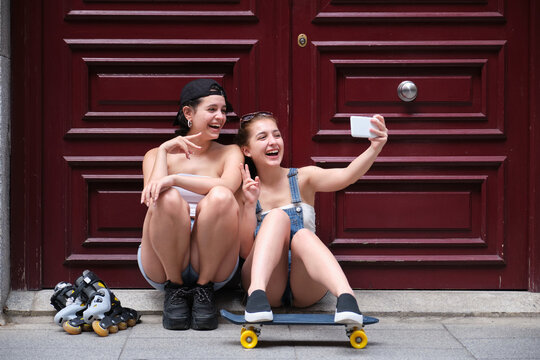 Two women laughing and taking a selfie, holding a skateboard and inline skates in a city. Friends, urban life.