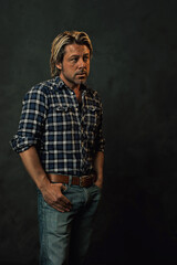 Man with blond hair in lumberjack shirt and jeans standing in front of a dark wall.