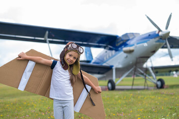 A little girl plays a pilot on the background of a small plane with a propeller. A child in a suit with cardboard wings dreams of flying