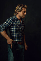 Man with blond hair in lumberjack shirt and jeans is holding a shotgun. Side view.