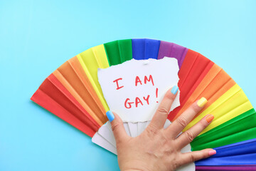 Text I am Gay on paper in hand with fake nails. Rainbow fan in colors of the rainbow, LGBTQ community pride symbol. Flat lay, top view on mint green, turquoise paper background.