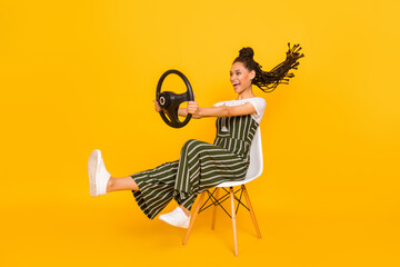 Full body photo of excited dark skin young woman sit chair hold steering wheel isolated on yellow color background