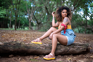Mulher negra de cabelo cacheado sentada em tronco de √°rvore no ch√£o em parque com folhas secas no ch√£o