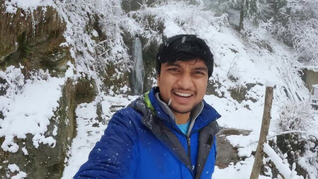 Selfie video of an Indian influencer speaking at the camera during snowfall at Jana waterfall in Manali, Himachal Pradesh, India. Snow sticks to the camera during snowfall while vlogging.