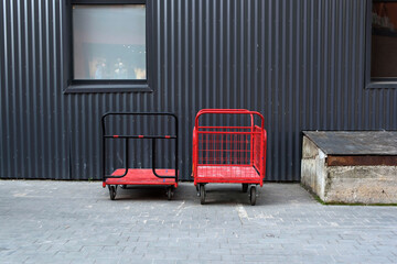 Metal red carts on the background of the grey facade of the warehouse. Corrugated gray metal facade. Manual transportation of goods. Cargo logistics. 