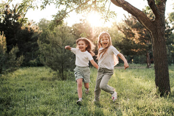 Naklejka na ściany i meble Happy cheerful sister child girls running in park together on green grass lawn outdoors. Happiness. Childhood. Togetherness. Summer season. Kids being themselves.
