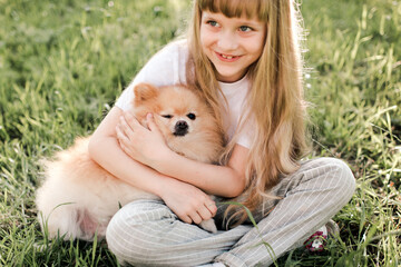 Pretty blonde kid girl 5-6 year old hold domestic pet sitting on green grass lawn outdoors in park. Looking at camera. Happiness. Friendship. Focus on dog.