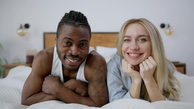 Portrait of happy joyful interracial couple posing lying on bed at home. Cheerful handsome African American tattooed man and beautiful blond Caucasian woman looking at camera resting in bedroom