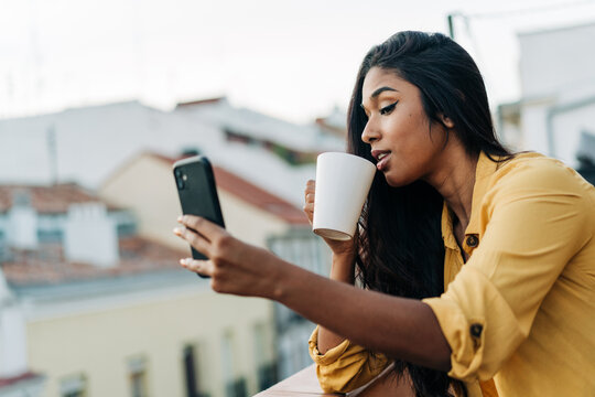 Hispanic woman drinking coffee and browsing smartphone on balcony