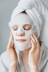 European woman applies white cloth moisturizing mask on her face, facial skin care and spa treatments at home, woman beautician makes anti-aging face mask, moisturizing and tightening mask, skin