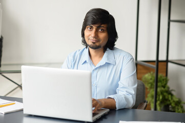 Handsome good-looking indian man in smart casual wear using laptop in contemporary office, mixed race eastern businessman looks at the camera. Technology and lifestyle concept