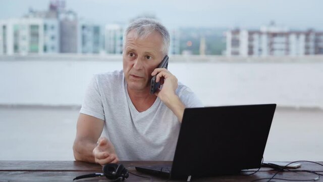An elderly white-haired businessman is seriously talking on the phone, sitting on a laptop in the evening on the balcony against the background of city lights in the summer.