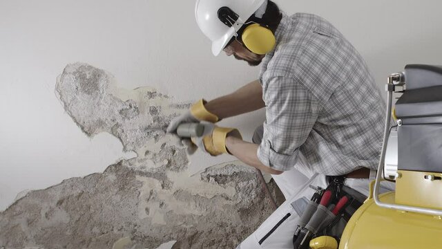 house renovation concept, construction worker wearing helmet at work breaks the old plaster of the wall with pneumatic air hammer chisel, with rubble and compressor on background