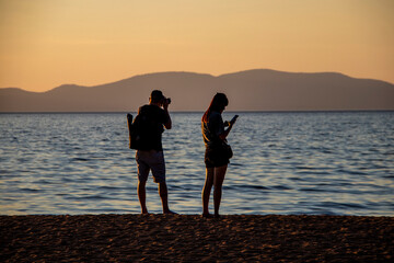 Young ouple in shorts  standing at beach - male photographing sunset and female looking at her phone - sihlouttes - Lake Tahoe USA.