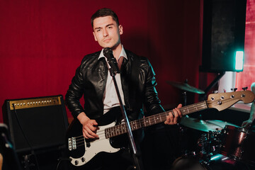 Talented handsome young guitarist man singing a song in studio recording on red background surrounded by instruments. Passion, hobby, singer, electric guitar