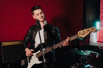 Talented handsome young guitarist man singing a song in studio recording on red background surrounded by instruments. Passion, hobby, singer, electric guitar