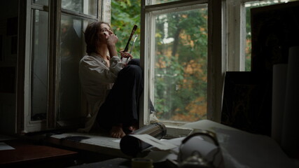 Serious woman smoking indoors. Contemporary painter relaxing on windowsill.