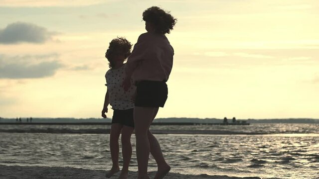 Slow motion video of happy young mother dancing and jumping with her teen daughter on baech during sunset