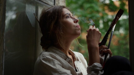 Tired painter smoking on windowsill. Young female artist resting in art studio.