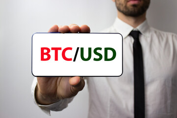 The bitcoin and dollar symbols are green and red on a white background of a mobile phone in the hand of a man with a tie and a white shirt.
